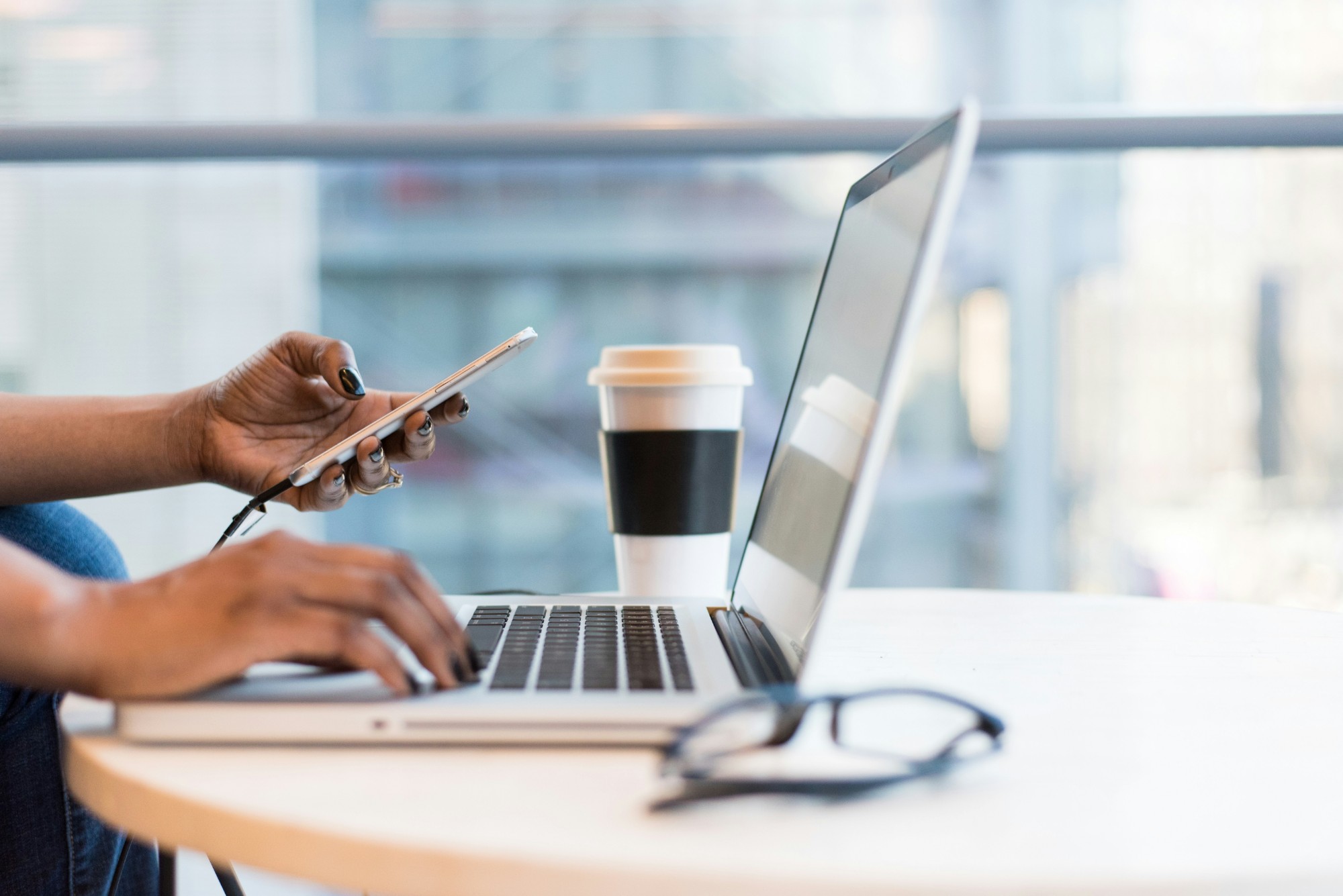Hands typing on a laptop beside a phone, coffee and glasses